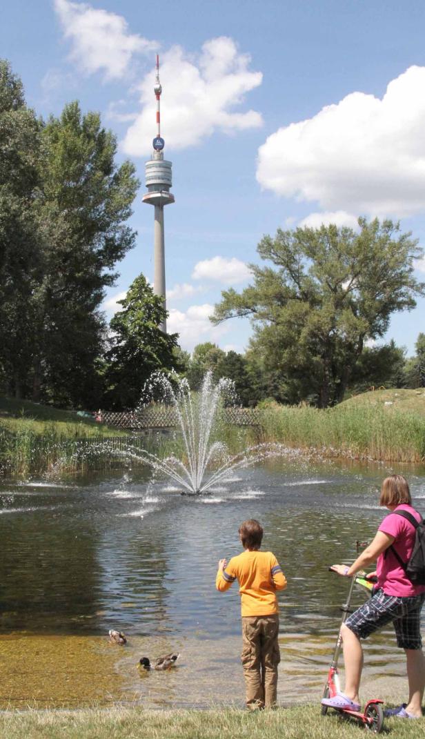 Ein Junge und ein Mädchen mit Roller beobachten Enten in einem Teich mit Springbrunnen und dem Donauturm im Hintergrund.
