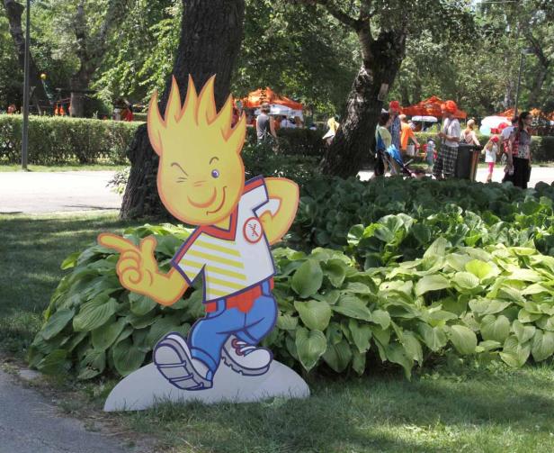 Eine Pappfigur eines gelben Maskottchens mit einem Augenzwinkern in einem Park.