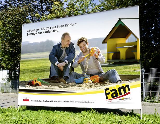Ein Werbeplakat zeigt zwei Männer, die in einem Sandkasten mit Spielzeug spielen.
