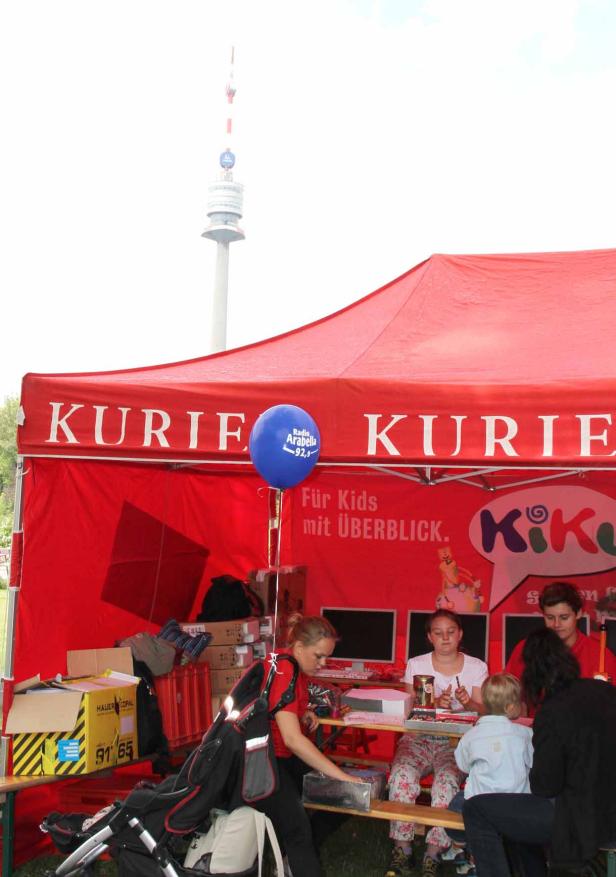 Kinder sitzen an einem Stand unter einem roten Zelt mit der Aufschrift „Kurier“ und basteln. Im Hintergrund ist der Donauturm zu sehen.