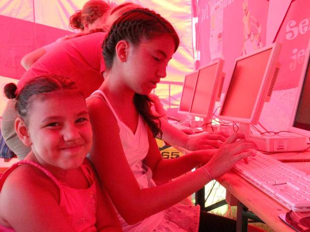 Zwei Mädchen sitzen an Computern, eines tippt auf der Tastatur, das andere lächelt in die Kamera.