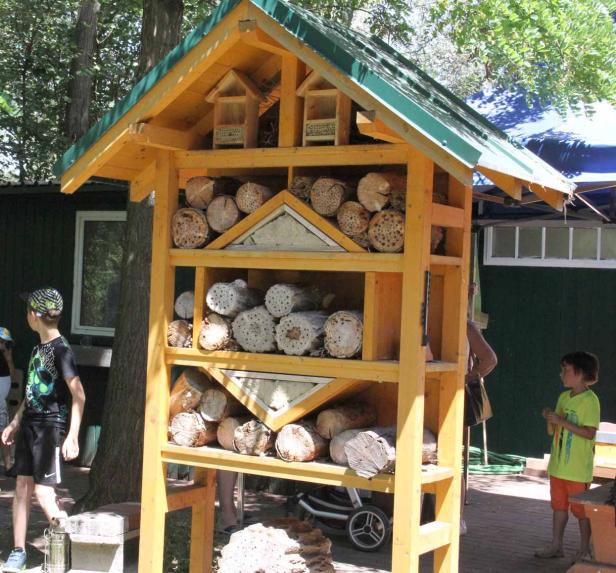 Ein gelbes Insektenhotel steht im Garten, gefüllt mit Holzstücken und kleinen Häuschen.