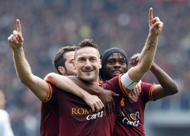 Francesco Totti wird von zwei Teamkollegen umarmt und jubelt mit erhobenem Zeigefinger.
