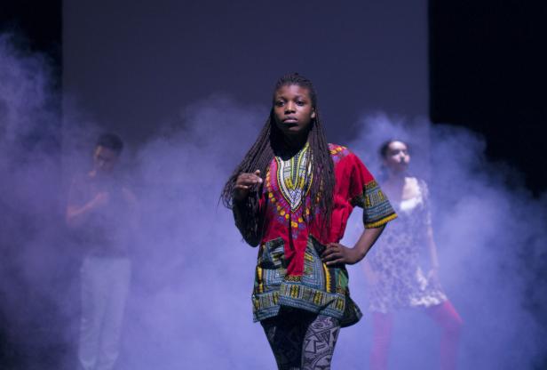 Eine junge Frau in einem farbenfrohen afrikanischen Hemd steht auf einer Bühne mit zwei Tänzern im Hintergrund.