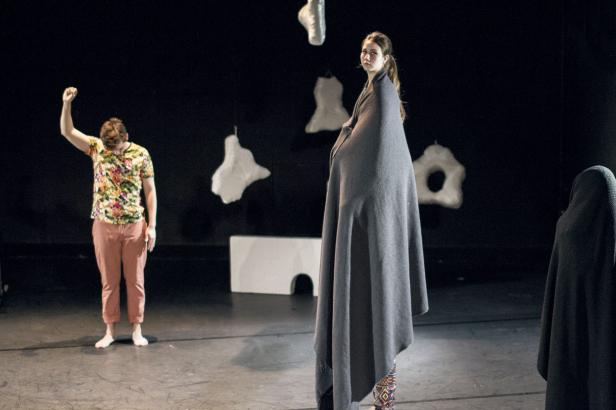 Auf einer Bühne stehen zwei in Decken gehüllte Personen und ein Mann mit erhobener Faust.