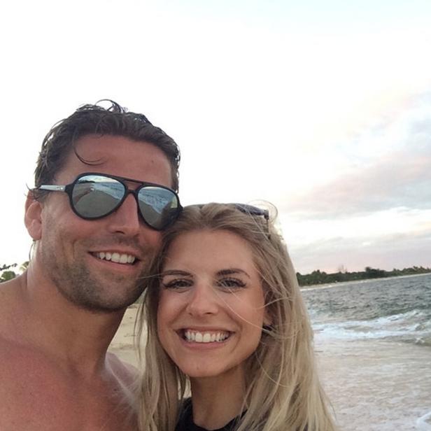 Ein Mann mit Sonnenbrille und eine Frau lächeln am Strand in die Kamera.