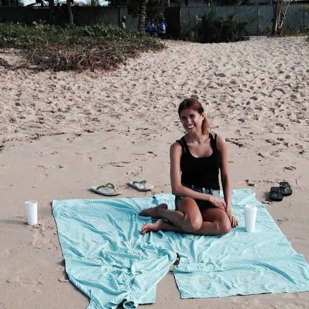 Eine junge Frau sitzt auf einem hellblauen Handtuch am Strand.