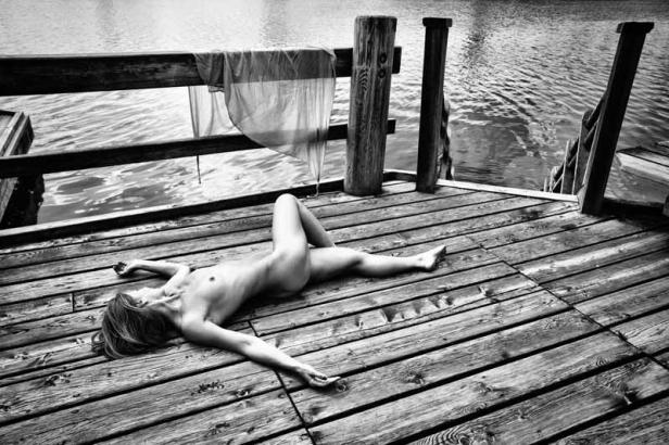Eine Frau liegt nackt auf einem Holzsteg am Wasser.