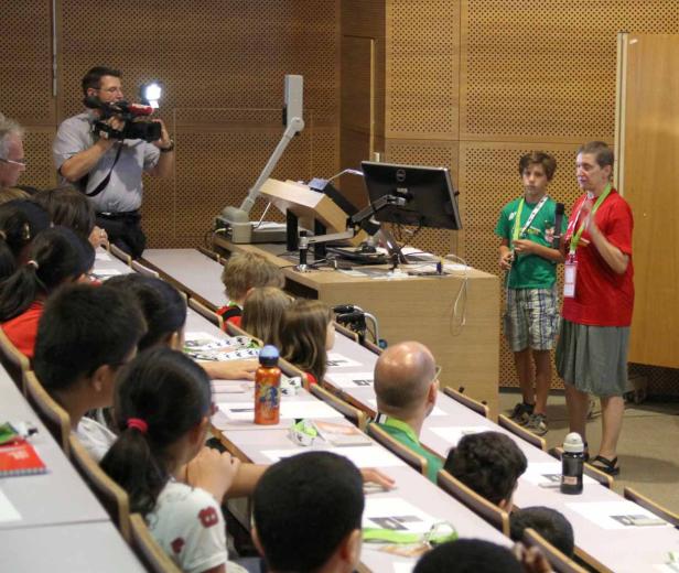 Ein Vortragssaal mit Kindern, einem Kameramann und zwei Personen vor einem Tisch mit einem Monitor.