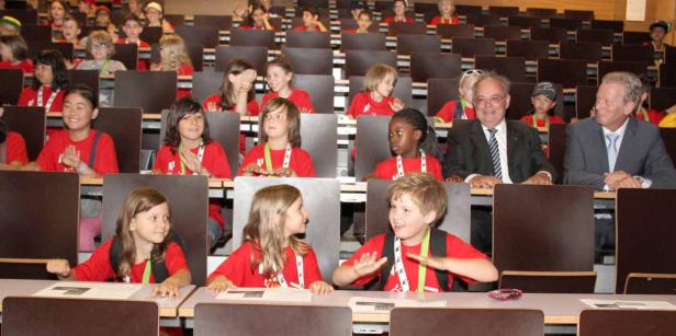 Kinder sitzen mit zwei Männern in einem Hörsaal.