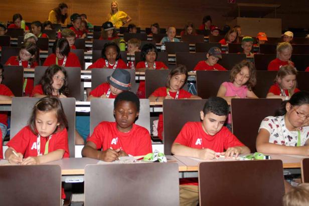 Eine Gruppe Kinder sitzt in einem Hörsaal und schreibt oder zeichnet.