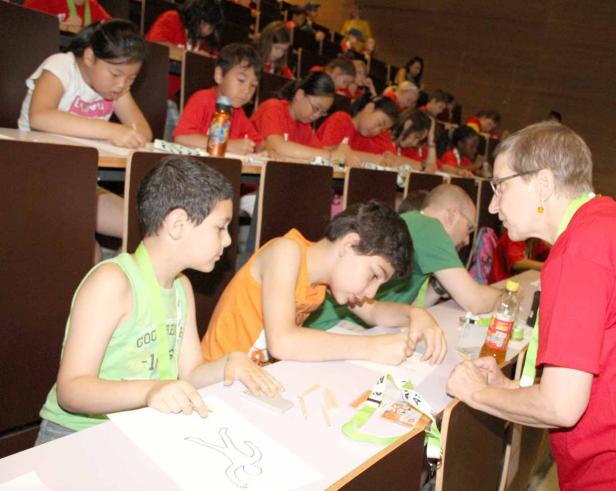 Kinder sitzen in einem Hörsaal und basteln mit Papier, während eine Frau ihnen hilft.