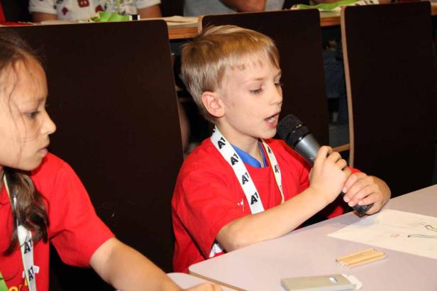 Ein Junge spricht in ein Mikrofon, während ein Mädchen daneben sitzt.