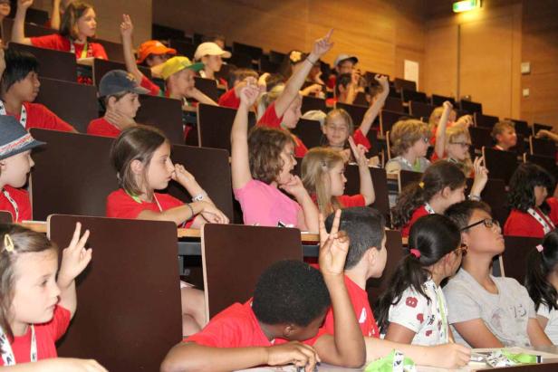 Eine Gruppe Kinder sitzt in einem Hörsaal und meldet sich.
