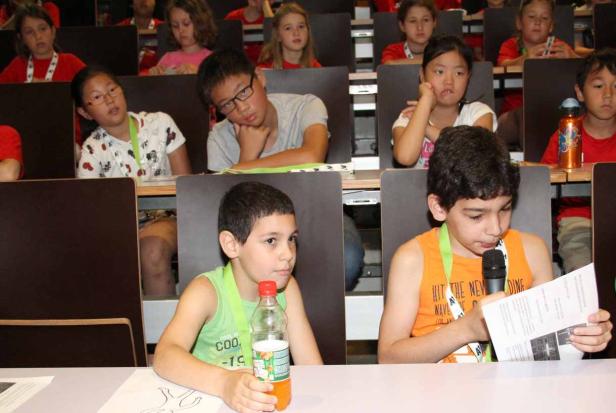 Kinder sitzen in einem Hörsaal, ein Junge spricht ins Mikrofon.