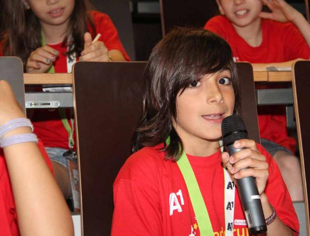 Ein Kind spricht in ein Mikrofon, während andere Kinder im Hintergrund sitzen.