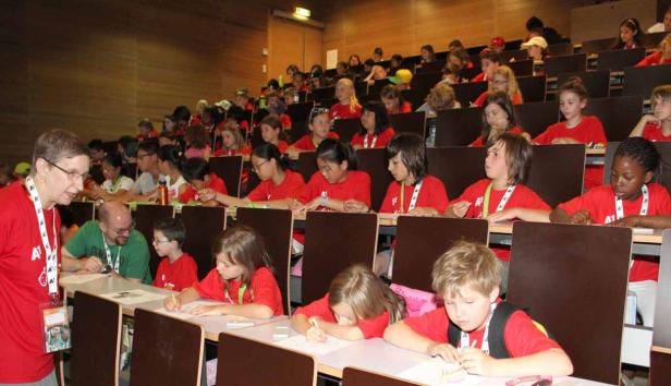 Kinder sitzen in einem Hörsaal und arbeiten an ihren Tischen, während eine Frau sie beaufsichtigt.