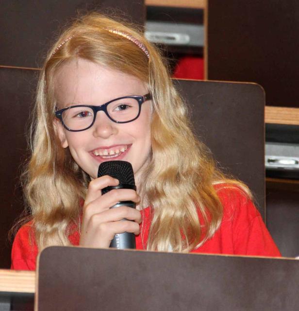 Ein blondes Mädchen mit Brille lächelt und hält ein Mikrofon in der Hand.