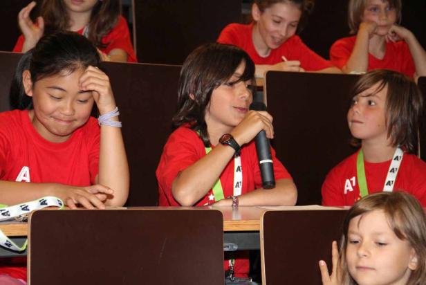 Eine Gruppe von Kindern sitzt in einem Raum, eines spricht in ein Mikrofon.
