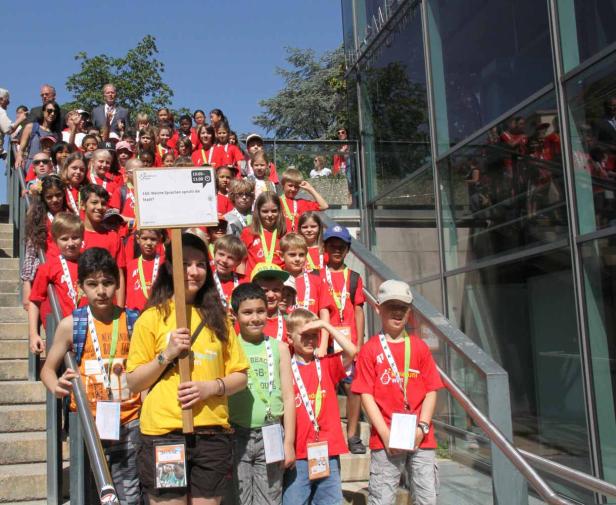 Eine Gruppe Kinder steht auf einer Treppe und hält ein Schild mit der Aufschrift „160: Welche Sprachen spricht die Stadt?“.