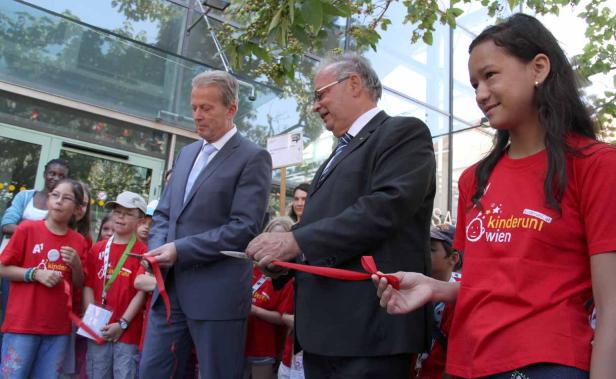 Zwei Männer im Anzug durchschneiden ein rotes Band, umgeben von Kindern.