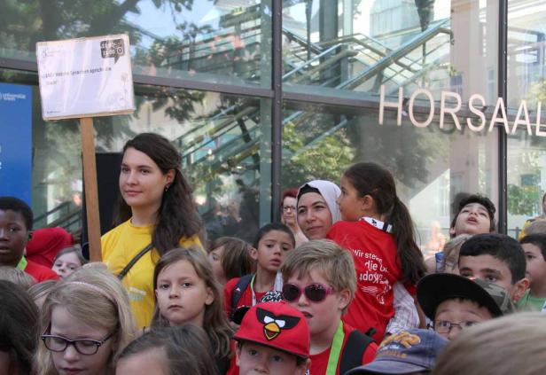 Eine Frau hält ein Schild mit der Aufschrift „160: Welche Sprachen spricht die Stadt?“ vor einer Gruppe von Kindern.