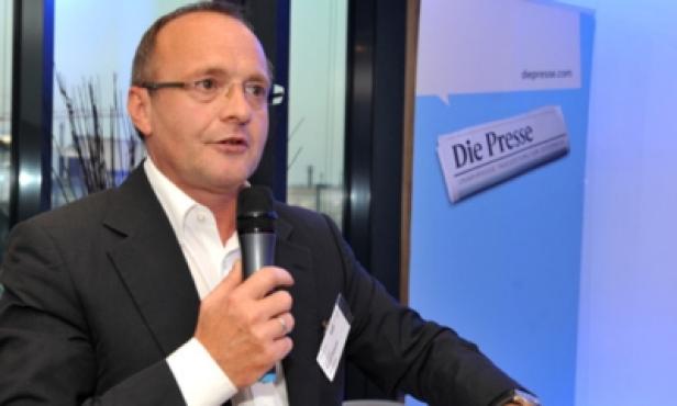 Ein Mann mit Brille spricht in ein Mikrofon vor einem Banner von „Die Presse“.