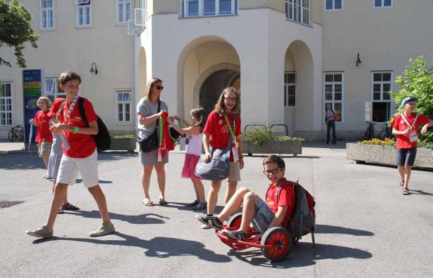 Eine Gruppe von Kindern und eine Frau sind vor einem Gebäude unterwegs, ein Junge sitzt auf einem Pedal-Gokart.