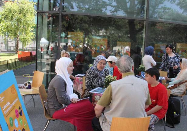 Eine Gruppe von Menschen sitzt an Tischen im Freien vor einem Gebäude.