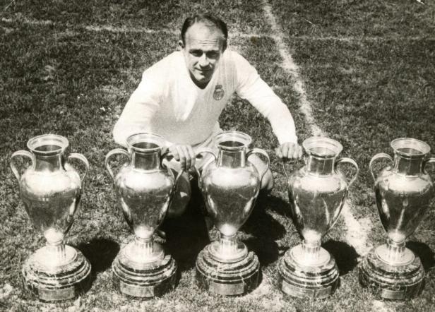 Alfredo Di Stéfano kniet mit fünf Europapokal-Trophäen auf einem Fußballfeld.