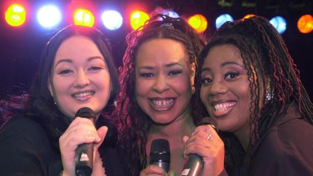 Drei Sängerinnen stehen auf der Bühne und singen in Mikrofone.