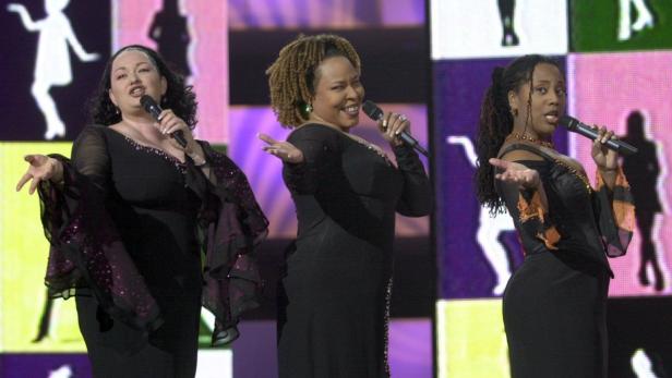 Drei Sängerinnen stehen auf einer Bühne und singen in Mikrofone.