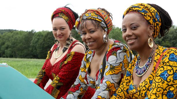 Drei Frauen mit farbenfrohen afrikanischen Kleidern und Kopfbedeckungen lächeln in die Kamera.