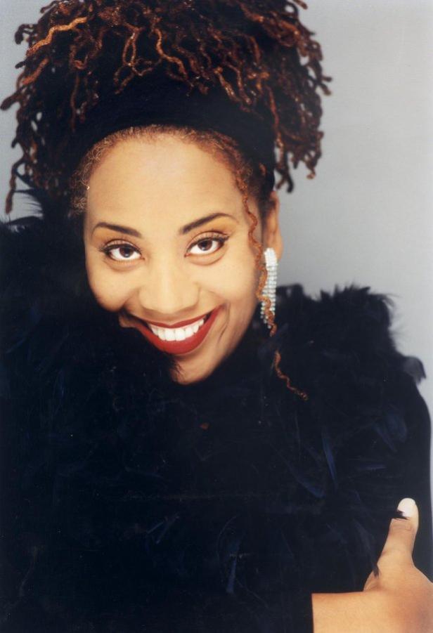 Eine lächelnde Frau mit Dreadlocks, dunklem Oberteil und auffälligen Ohrringen.