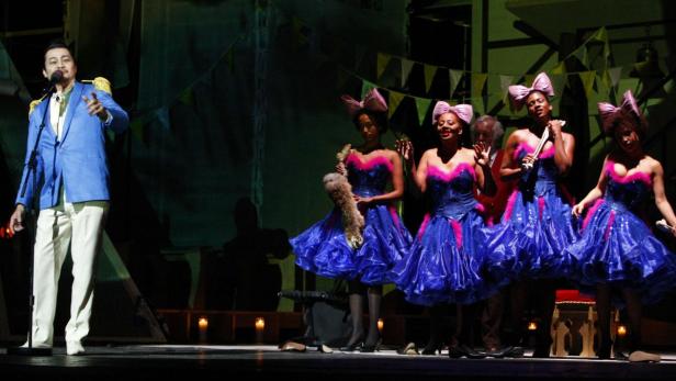 Ein Mann in einem blauen Anzug singt vor einer Gruppe von Tänzerinnen in blauen Kleidern auf einer Bühne.