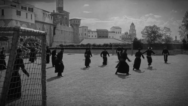 In einem Schwarzweißbild spielen Priester auf einem Rasenplatz Fußball.