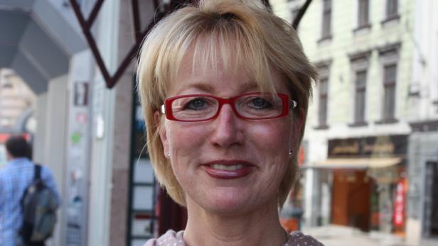 Eine lächelnde Frau mit blonden Haaren und roter Brille steht im Freien.