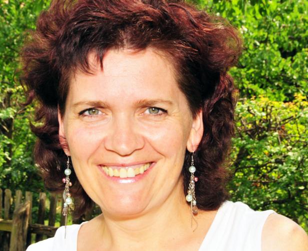 Eine lächelnde Frau mit roten Haaren und Ohrringen steht vor einem grünen Hintergrund.