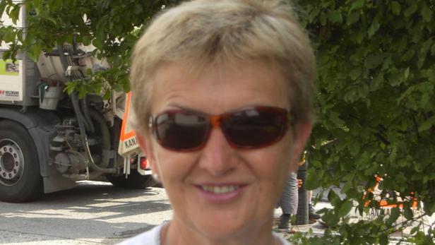 Eine lächelnde Frau mit Sonnenbrille vor einem Müllwagen und Bäumen.