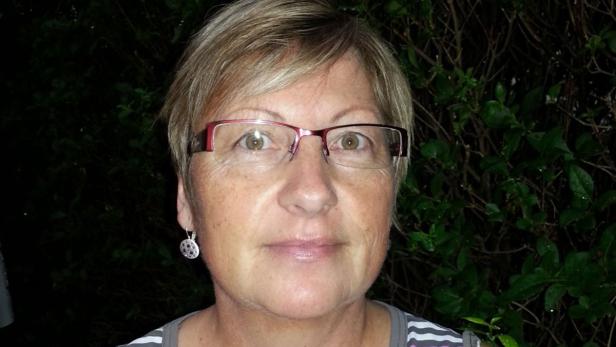 Eine Frau mit Brille und kurzem blonden Haar vor einem dunklen Hintergrund mit Pflanzen.