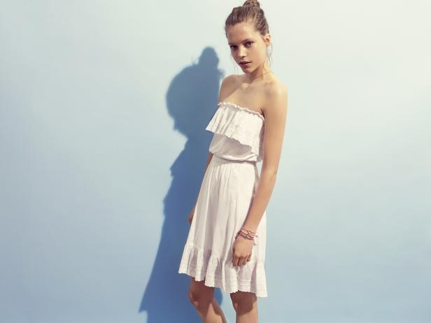 Eine junge Frau posiert in einem weißen Sommerkleid vor einem hellblauen Hintergrund.