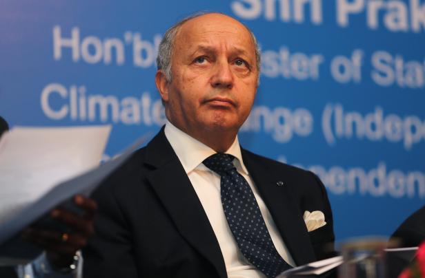 Ein Mann im Anzug vor einem blauen Hintergrund mit Text betrachtet etwas abseits der Kamera.