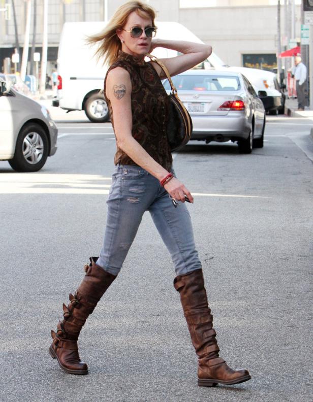 Eine Frau mit Sonnenbrille, braunen Stiefeln und Tätowierung geht über eine Straße.