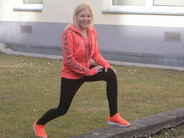 Eine blonde Frau dehnt sich in Sportkleidung auf einer Rasenfläche.