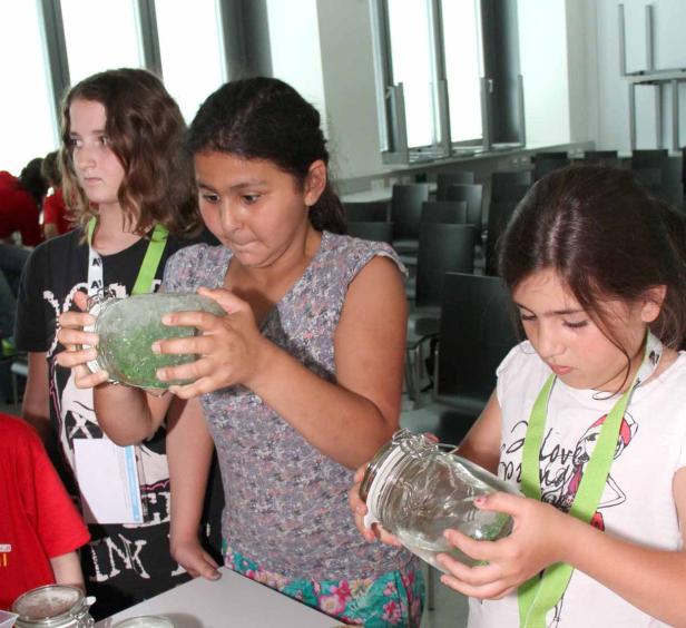Drei Mädchen betrachten Gläser mit Flüssigkeiten in einem hellen Raum.