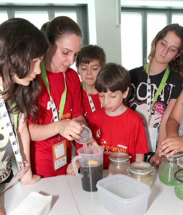 Eine Frau gießt mit einem Sieb Flüssigkeit in einen Messbecher, umgeben von Kindern.