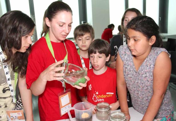 Eine Frau zeigt einer Gruppe von Kindern ein Glas mit grünen Kügelchen.