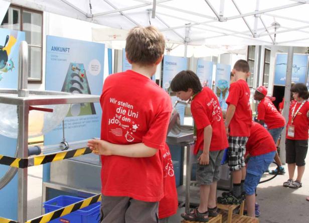 Eine Gruppe Kinder in roten T-Shirts betrachtet eine Wasserinstallation im Freien.