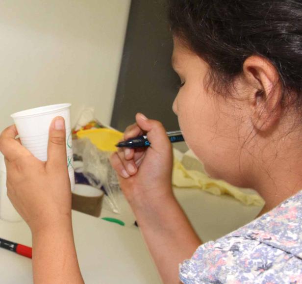 Ein Mädchen bemalt einen Plastikbecher mit einem Stift.