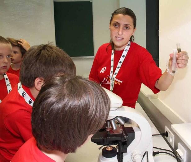 Eine Frau erklärt Kindern etwas über ein Mikroskop und hält Reagenzgläser in der Hand.